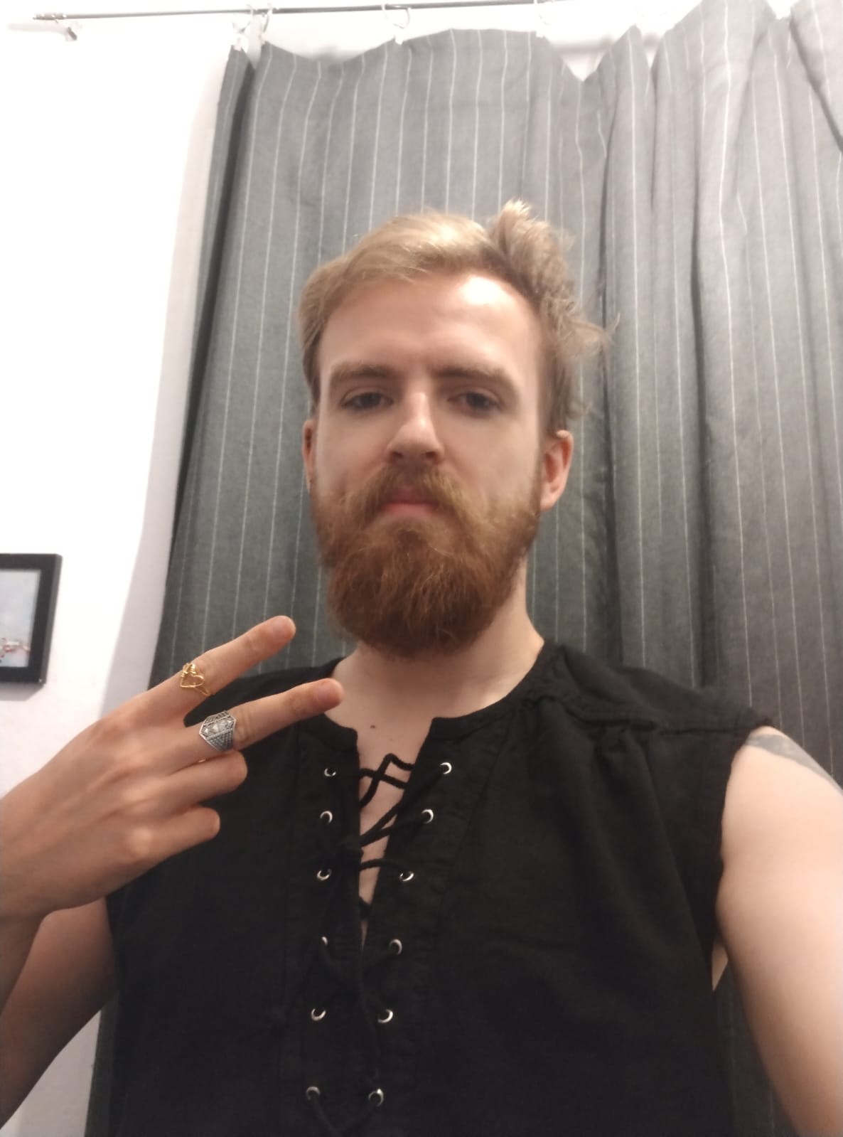 blond beared person in a goth tanktop showing a hand with peace at the camera
